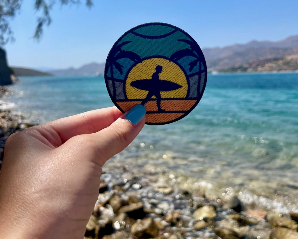 Frauenhand hält den Surfer Patch mit dem Meer im Hintergrund