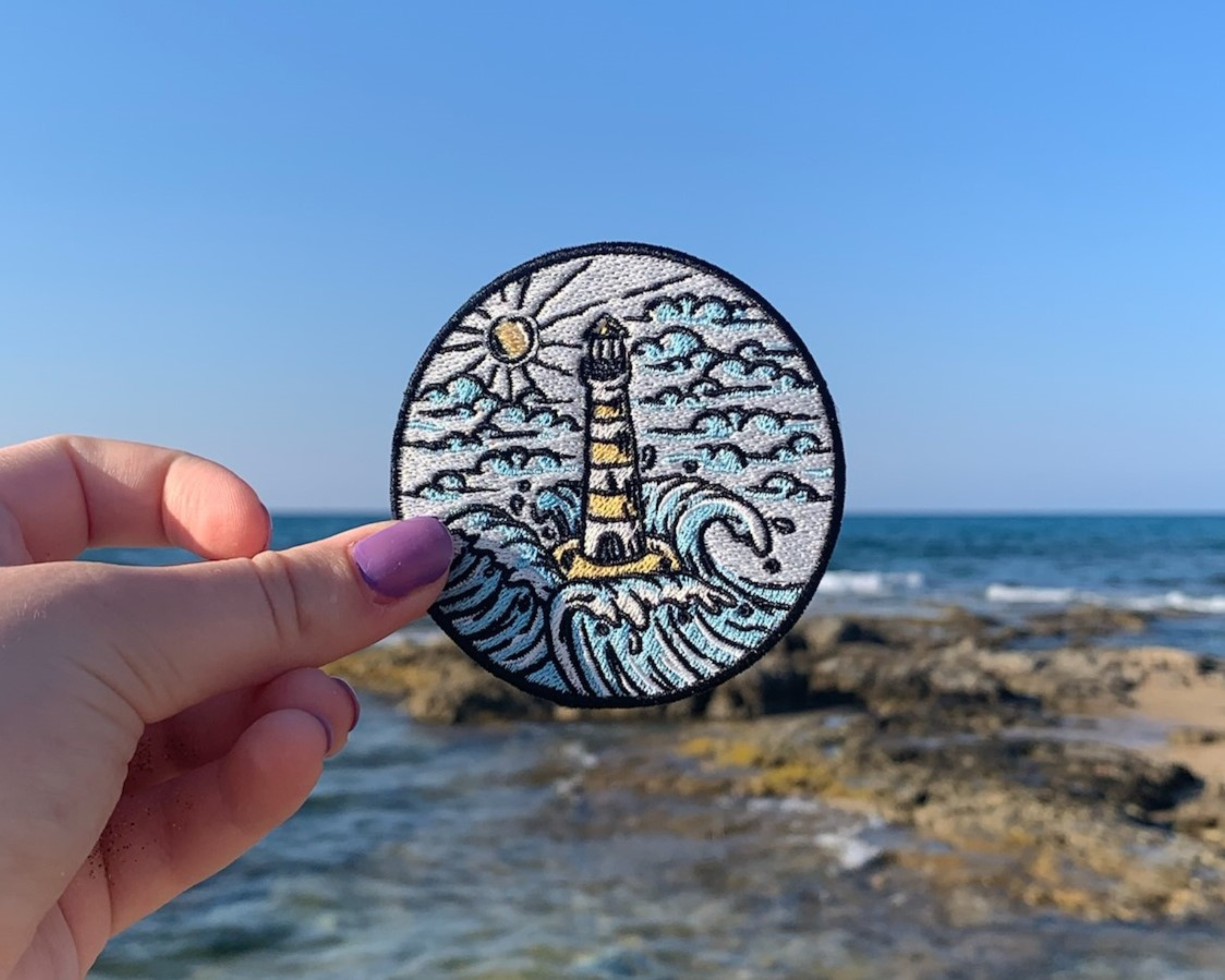 Frauenhand hält den Leuchtturm Patch mit dem Meer im Hintergrund