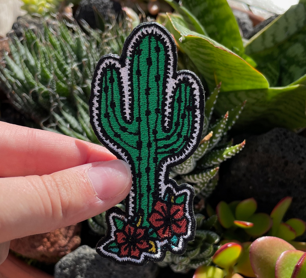 Kaktus Blumen Patch gehalten von einer Hand mit Kakteen im Hintergrund