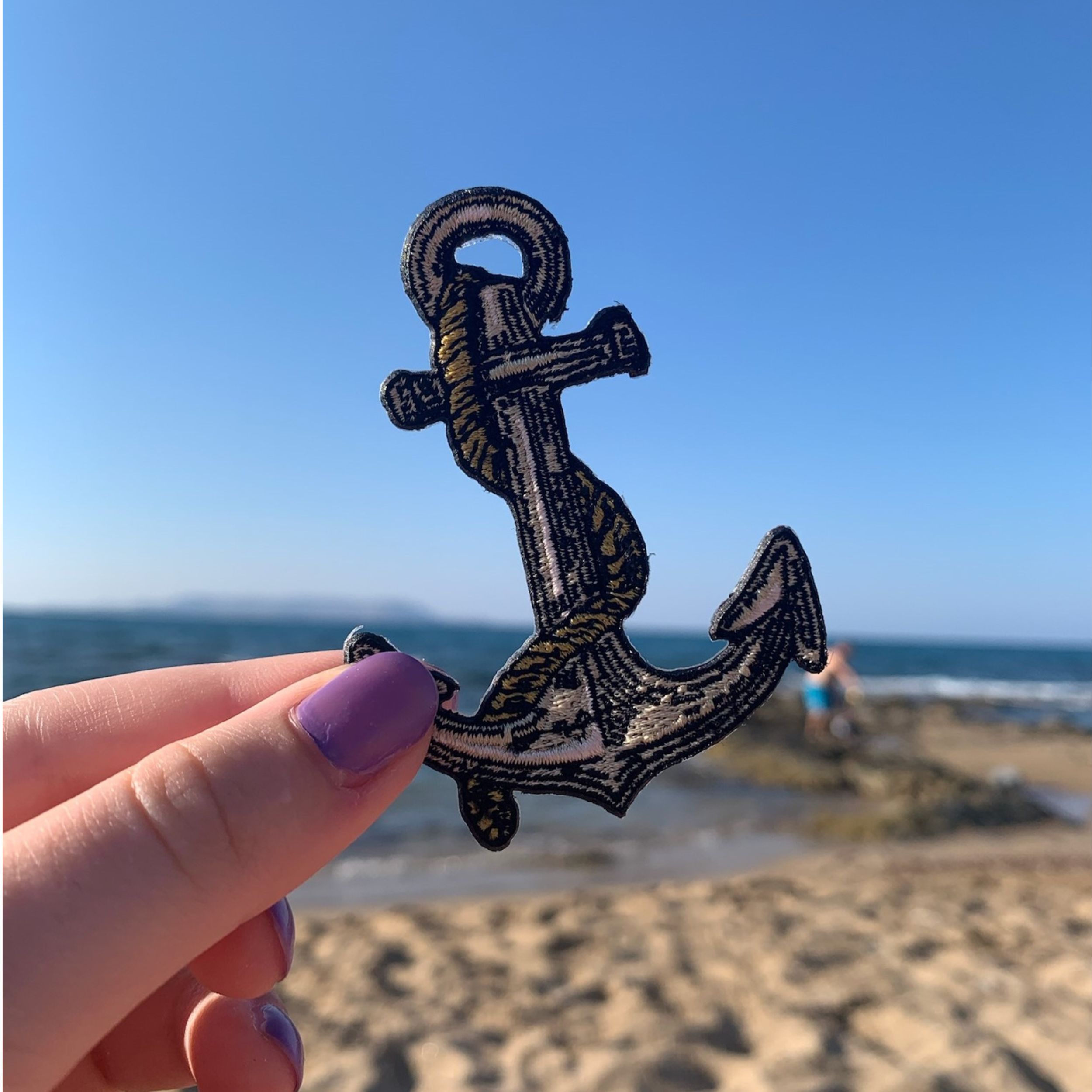 Frauenhand hält den Anker Patch im Urlaub mit Strand im Hintergrund