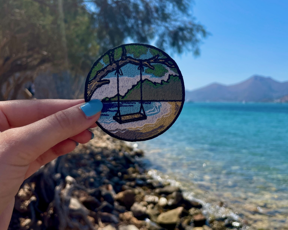 Strand Schaukel Patch mit Meer  und Steinen im Hintergund gehalten von einer Frauenhand